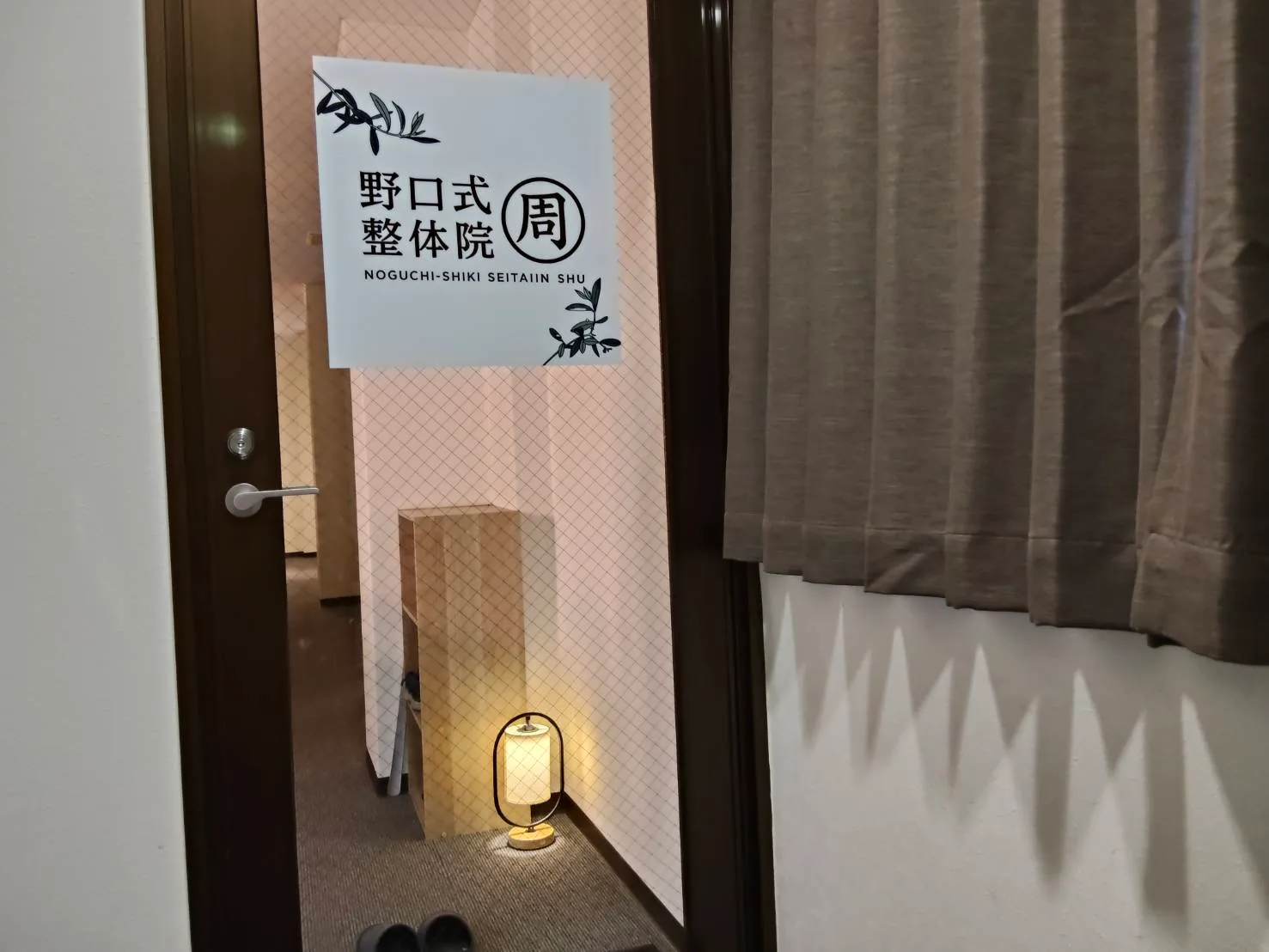 院の入口。白い看板に「野口式整体院 周」のロゴが掲げられ、足元に和紙の行灯が灯っている